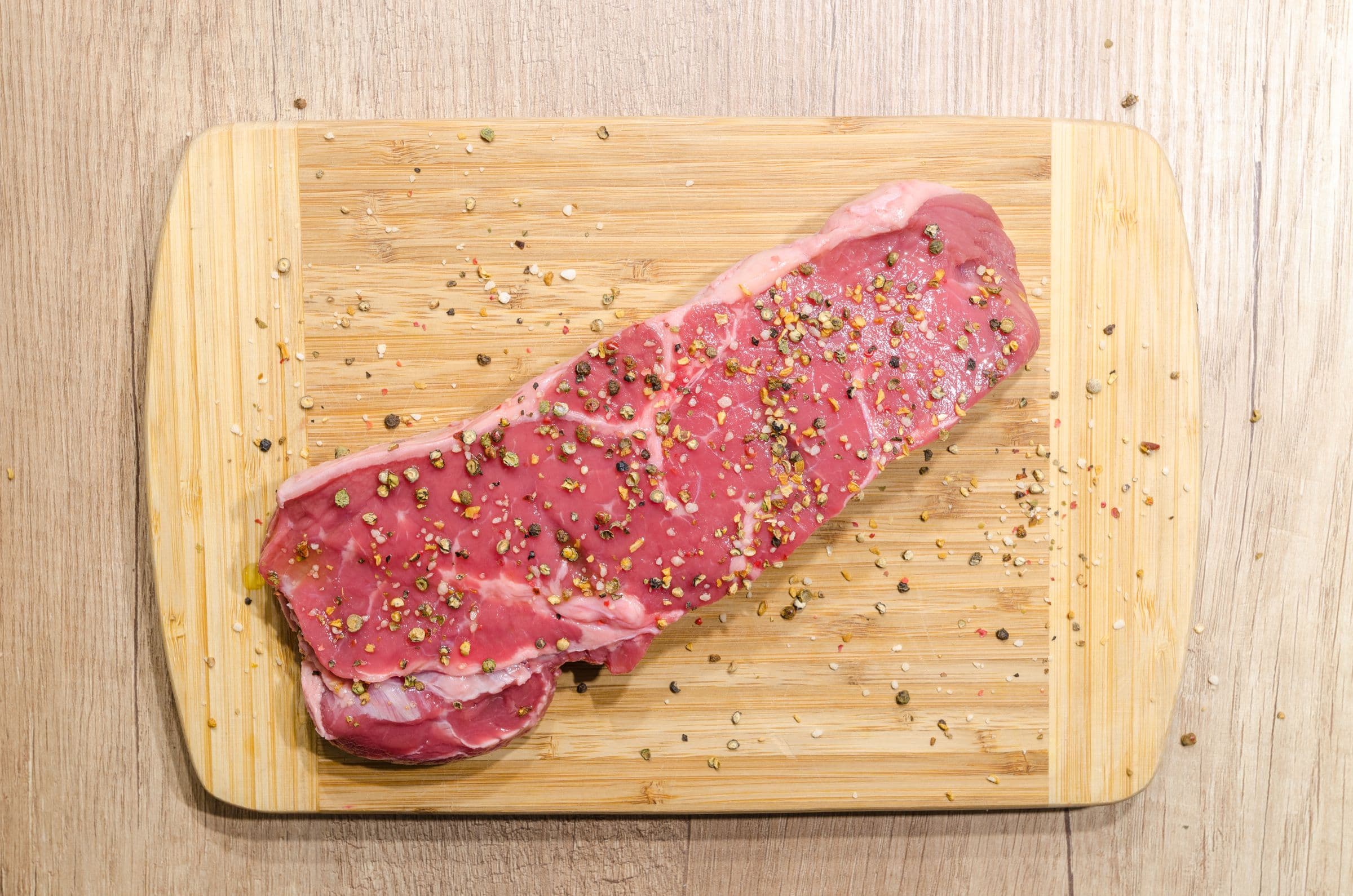 Raw beef steak on a wooden board, seasoned with peppercorns and salt